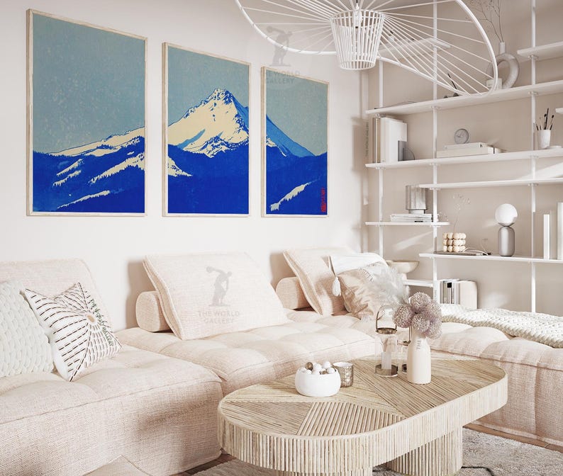 K&ouml;nnte beinhalten: Drei gerahmte Kunstdrucke, die eine Bergkette in Blau- und Wei&szlig;t&ouml;nen zeigen. Das Kunstwerk h&auml;ngt &uuml;ber einem hellfarbigen Ecksofa mit dekorativen Kissen. Ein Holztisch und ein wei&szlig;es Regal vervollst&auml;ndigen die Einrichtung des Raumes.