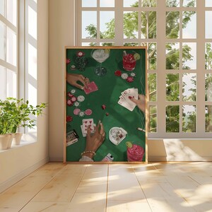 May include: A framed art print depicts a poker game scene on a green felt table. Hands are holding playing cards, poker chips, and cocktails. The artwork is set against a bright window and wooden floor.