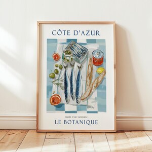 May include: A framed art print featuring a still life of sardines, olives, and bread on a checkered tablecloth. The print includes the text "C&Ocirc;TE D'AZUR" and "LE BOTANIQUE". The color palette includes blue, white, red, and green.