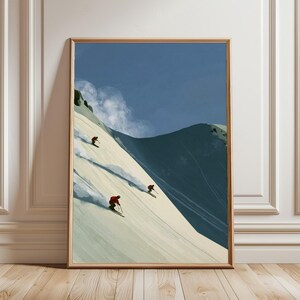 May include: A framed vertical art print depicting three skiers in red jackets descending a steep, snow-covered mountain slope under a deep blue sky. The scene features a large, dark mountain peak in the background and soft, white clouds.