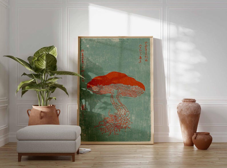 May include: A framed art print featuring a large red mushroom against a teal background. The artwork has Japanese text and is displayed in a room with a potted plant, a gray ottoman, and decorative pottery.