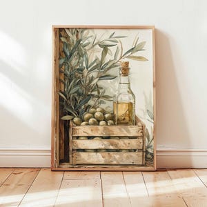 Op de afbeelding: Een ingelijste aquarelprint met een houten kist vol groene olijven. Een heldere glazen fles met een kurk bevat olijfolie. Olijftakken en -bladeren creëren een rustiek, natuurlijk thema.