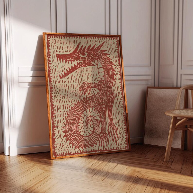 May include: A framed print featuring a red dragon illustration on a cream background. The dragon is depicted with sharp teeth and a long, coiling tail. The artwork is set in a wooden frame and leans against a white wall.