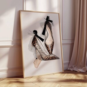 Op de afbeelding: Waterverfprint van twee hoge hakken. E&eacute;n schoen heeft een luipaardprint, de andere is effen van kleur. Beide schoenen hebben zwarte strikken aan de bovenkant. Het kunstwerk is ingelijst in een licht houten frame en leunt tegen een muur.