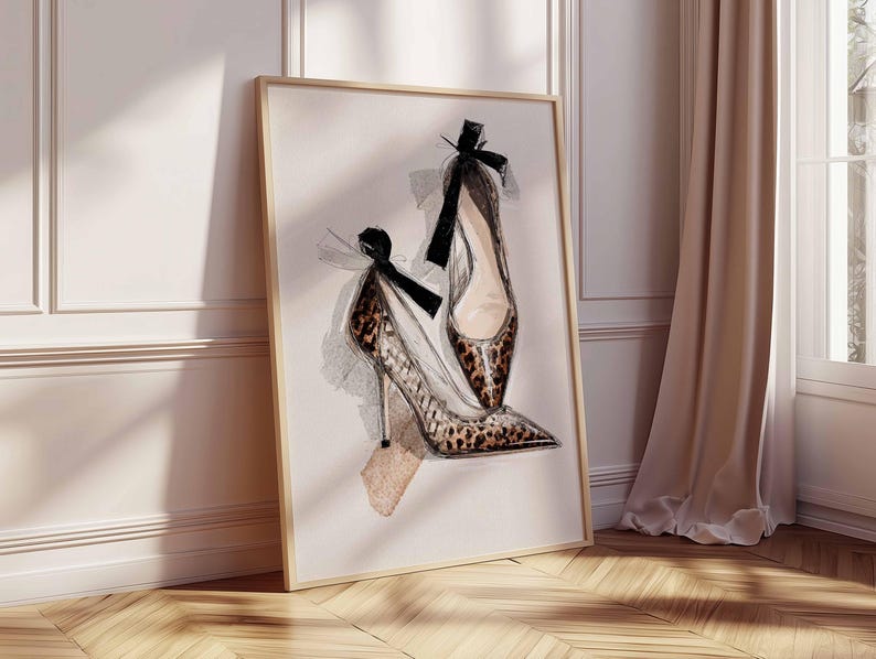Op de afbeelding: Waterverfprint van twee hoge hakken. E&eacute;n schoen heeft een luipaardprint, de andere is effen van kleur. Beide schoenen hebben zwarte strikken aan de bovenkant. Het kunstwerk is ingelijst in een licht houten frame en leunt tegen een muur.