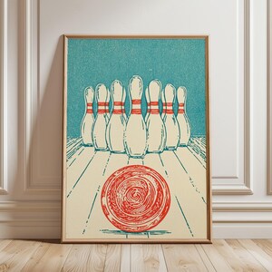 May include: A framed art print featuring a stylised illustration of ten white bowling pins with red stripes arranged in a triangle formation on a light cream lane, with a large, textured red bowling ball rolling towards them on a teal background.