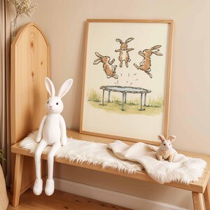 May include: A wooden bench with a white plush bunny and a framed print of rabbits jumping on a trampoline. A smaller plush bunny sits on a white blanket. The artwork and plush toys create a whimsical, playful theme.