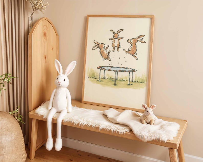May include: A wooden bench with a white plush bunny and a framed print of rabbits jumping on a trampoline. A smaller plush bunny sits on a white blanket. The artwork and plush toys create a whimsical, playful theme.