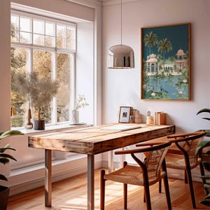 May include: A bright, airy home office with a large wooden desk and two woven chairs. A framed artwork depicts a serene landscape with a lake, palm trees, and ornate buildings. A large window offers a view of trees.
