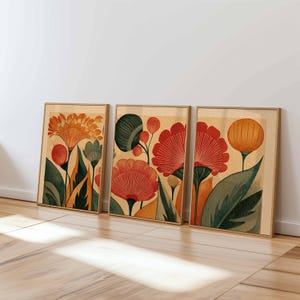 Pode incluir: Três impressões de arte floral emolduradas. Cada impressão apresenta flores estilizadas em tons de laranja, vermelho e verde sobre um fundo bege. As molduras são de cor de madeira clara e a obra de arte está encostada a uma parede branca.