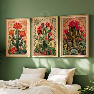 May include: Three framed botanical prints featuring illustrations of cacti with red and pink flowers. The prints are set against a green wall and above a bed with white linens.