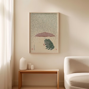 May include: A framed print depicting a green frog sheltering under a red umbrella in the rain. The artwork is in the style of a Japanese woodblock print, with a light wood frame. The frog holds the umbrella, and rain falls in the background.