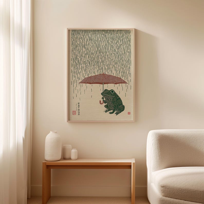 May include: A framed print depicting a green frog sheltering under a red umbrella in the rain. The artwork is in the style of a Japanese woodblock print, with a light wood frame. The frog holds the umbrella, and rain falls in the background.