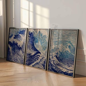 May include: Three framed art prints depicting ocean waves in shades of blue and beige. The artwork has a woodcut style, with detailed lines creating the wave patterns. The frames are black, and the prints are leaning against a white wall.