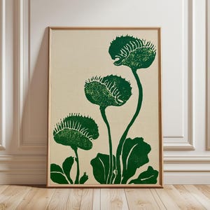 May include: A rectangular wall art print featuring a stylized illustration of three green Venus flytrap plants with textured leaves and spiky edges, set against a solid light beige background and enclosed in a simple light-colored wooden frame.