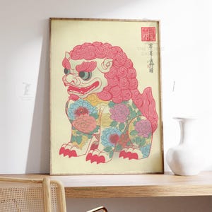 May include: Framed print of a stylized, seated lion-like creature with a pink mane and floral patterns. The artwork features a cream-colored background and traditional Japanese text and seals. The frame is a light brown color.