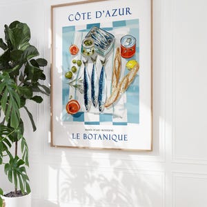 May include: A framed art print featuring a still life of French cuisine. The print includes the text "C&Ocirc;TE D'AZUR" and "MUS&Eacute;E D'ART MODERNE LE BOTANIQUE". The image depicts sardines, olives, bread, and citrus fruits on a blue and white checkered tablecloth.