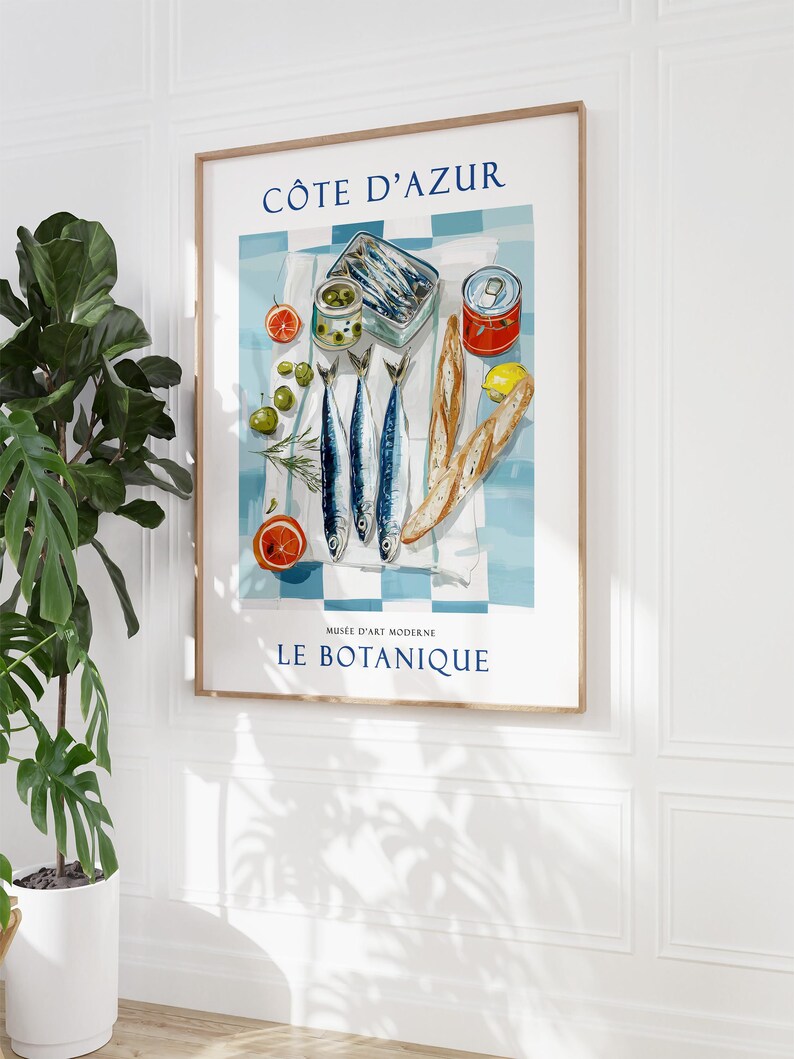May include: A framed art print featuring a still life of French cuisine. The print includes the text "C&Ocirc;TE D'AZUR" and "MUS&Eacute;E D'ART MODERNE LE BOTANIQUE". The image depicts sardines, olives, bread, and citrus fruits on a blue and white checkered tablecloth.