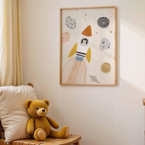 May include: A framed art print featuring a whimsical rocket ship with a cat in the cockpit, surrounded by planets and stars. The artwork is in a child's room, with a teddy bear on a wooden chair.