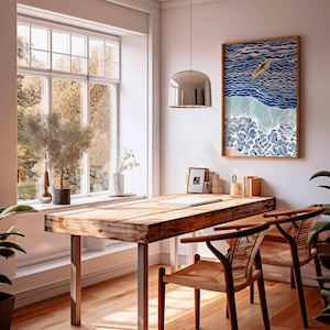 May include: A bright, airy room with a wooden desk and two chairs. A framed artwork depicting a surfer on waves hangs on the wall. A large window offers a view of trees. A silver pendant light hangs above the desk.
