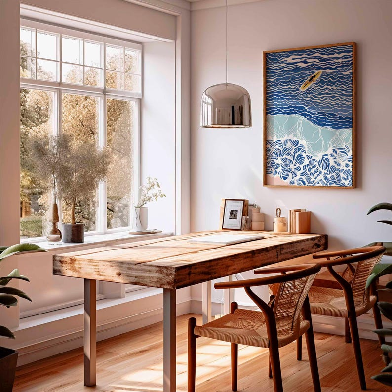 May include: A bright, airy room with a wooden desk and two chairs. A framed artwork depicting a surfer on waves hangs on the wall. A large window offers a view of trees. A silver pendant light hangs above the desk.