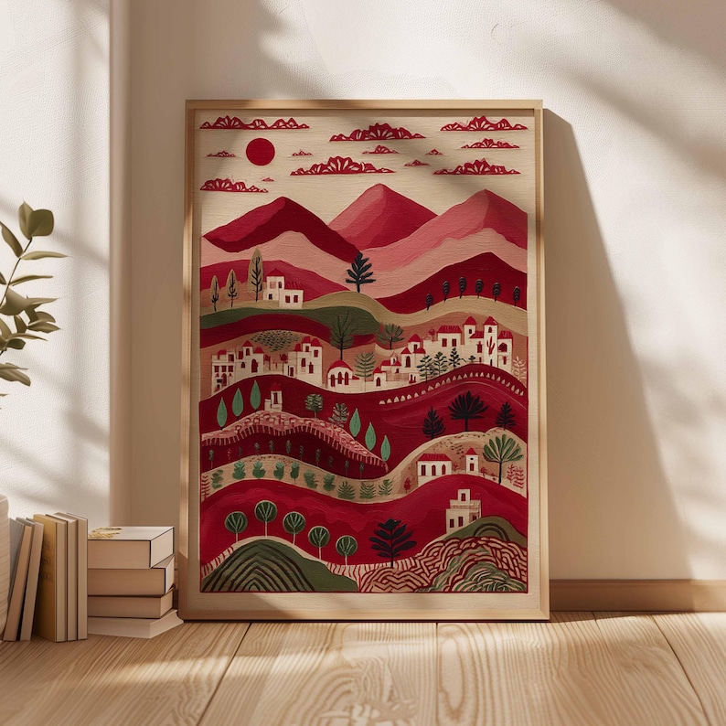 May include: Framed art print showing a stylized landscape. The artwork features rolling hills, mountains, and a village in shades of red, pink, and white, with a red sun and clouds. The print is in a light-coloured wooden frame.