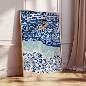 May include: Framed art print depicting a surfer on a yellow surfboard in the ocean. The artwork features a stylized illustration of ocean waves in shades of blue, with a light pink beach and a wooden frame. The art is leaning against a white wall and wooden floor.
