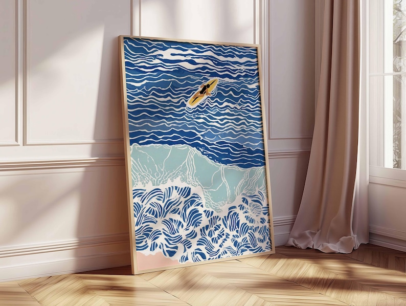 May include: Framed art print depicting a surfer on a yellow surfboard in the ocean. The artwork features a stylized illustration of ocean waves in shades of blue, with a light pink beach and a wooden frame. The art is leaning against a white wall and wooden floor.