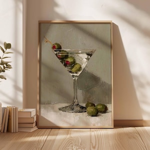 May include: A framed painting depicts a martini glass filled with a clear liquid and green olives. The glass sits on a white surface against a muted green background. The artwork is in a wooden frame.