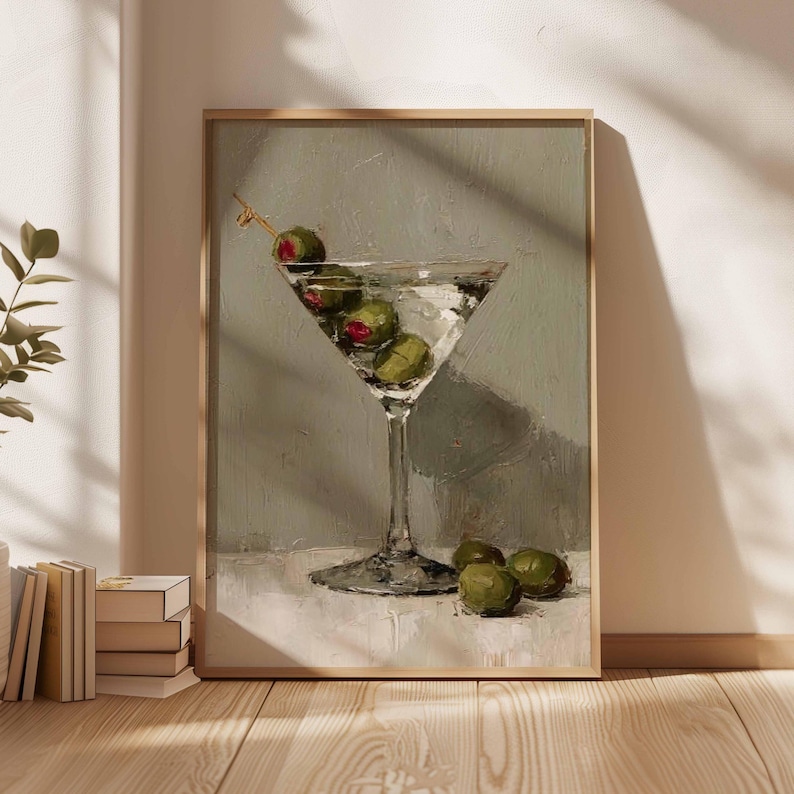 May include: A framed painting depicts a martini glass filled with a clear liquid and green olives. The glass sits on a white surface against a muted green background. The artwork is in a wooden frame.