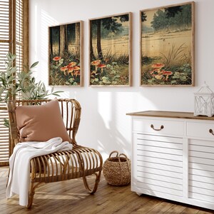 May include: Three framed art prints showing a forest scene with red-capped mushrooms. The artwork features trees, grass, and a pond, with a muted colour palette of greens, browns, and reds. The prints are displayed above a white cabinet and near a wicker chair.