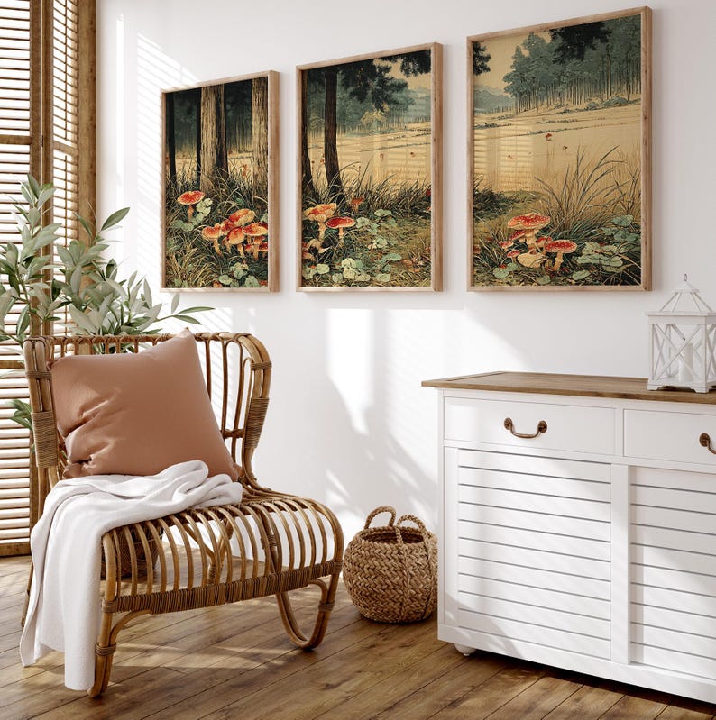 May include: Three framed art prints showing a forest scene with red-capped mushrooms. The artwork features trees, grass, and a pond, with a muted colour palette of greens, browns, and reds. The prints are displayed above a white cabinet and near a wicker chair.