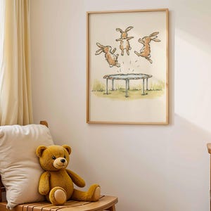 May include: A framed print featuring a whimsical illustration of three brown rabbits jumping on a trampoline. The artwork is set against a backdrop of a grassy field and a light-colored sky. The frame is made of light-colored wood. A teddy bear sits on a wooden chair.