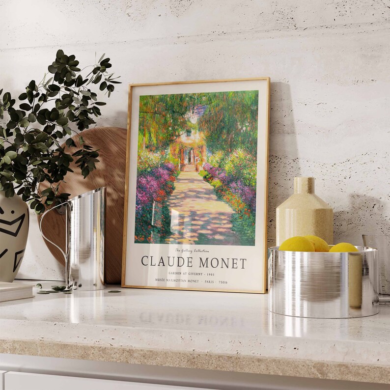 May include: A framed print of a painting by Claude Monet titled "Garden at Giverny - 1902". The painting depicts a path lined with flowers leading to a house in the distance. The print is on a white countertop with a silver pitcher, a wooden cutting board, and two silver bowls.