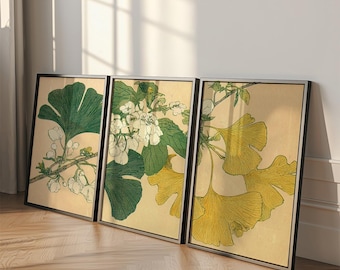 May include: Three framed botanical prints featuring detailed illustrations of ginkgo leaves and flowering branches. The artwork showcases a palette of green, yellow, and white against a beige background, with black frames. The prints are leaning against a wall.