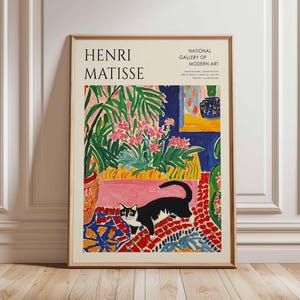 以下が含まれることがあります： アンリ・マティスのカラフルなインテリアシーンを描いた額入りのアートプリント。 白黒の猫が模様入りのラグの上にいて、鮮やかな植物や花に囲まれています。 上部には「HENRI MATISSE」の文字があります。