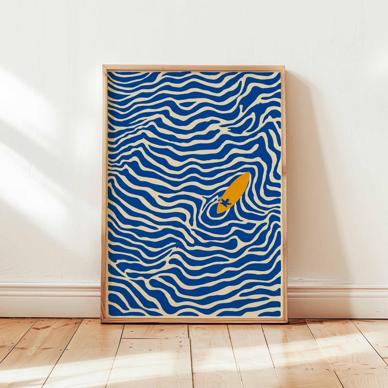 May include: A framed art print featuring a yellow surfboard with a surfer in blue and cream-colored wavy water. The artwork is in a wooden frame and leans against a white wall and wooden floor.