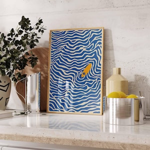 May include: A framed art print featuring a blue and white wavy pattern with a yellow surfboard in the center. The artwork is displayed on a white countertop with other decorative items.