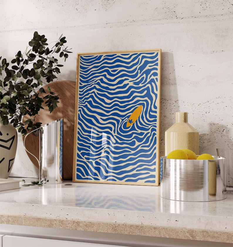 May include: A framed art print featuring a blue and white wavy pattern with a yellow surfboard in the center. The artwork is displayed on a white countertop with other decorative items.