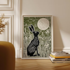May include: A framed art print depicting a black hare in a field of tall grass beneath a full moon. The background is a muted green, and the print is in a white frame. The artwork is displayed on a wooden floor.