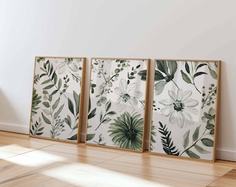 May include: Three framed botanical art prints. Each print features a watercolor design of green leaves and white flowers on a white background. The frames are a light brown color and are leaning against a white wall.
