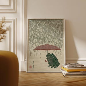 May include: A framed art print depicting a green frog sheltering under a red umbrella in the rain. The artwork has a Japanese style, with vertical lines representing rain and text in the lower left corner. The frog is detailed with textured patterns.