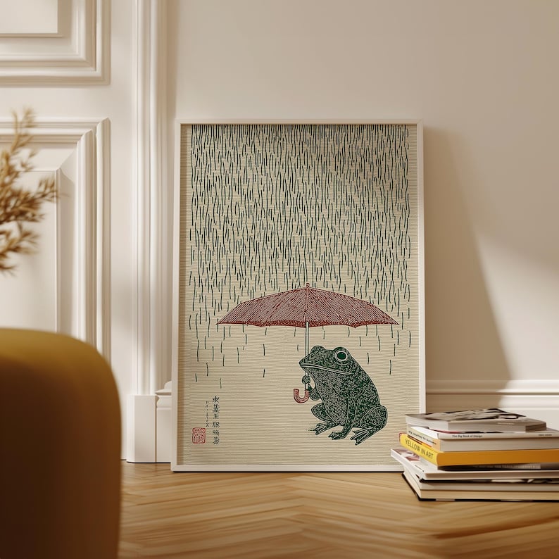 May include: A framed art print depicting a green frog sheltering under a red umbrella in the rain. The artwork has a Japanese style, with vertical lines representing rain and text in the lower left corner. The frog is detailed with textured patterns.