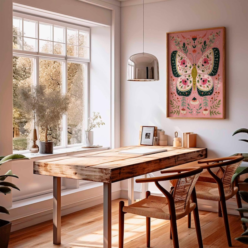 May include: A framed butterfly art print with a pink background hangs above a wooden desk. The butterfly design features green, pink, and white floral patterns. Two woven chairs sit at the desk. A large window and a silver pendant light are also visible.