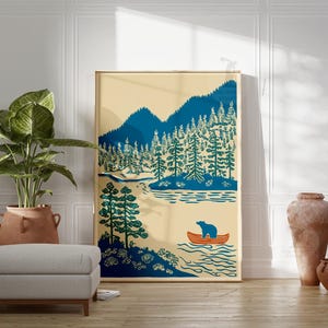 May include: A framed art print featuring a landscape scene with mountains, trees, and a river. A bear is in an orange canoe. The artwork uses shades of blue, green, and beige. The print is displayed in a room with a plant, ottoman, and decorative vases.