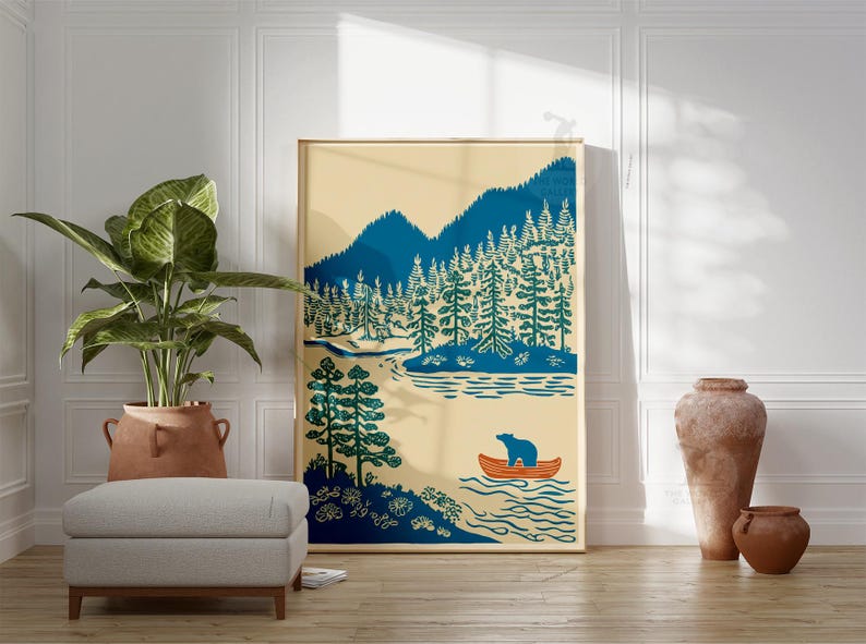 May include: A framed art print featuring a landscape scene with mountains, trees, and a river. A bear is in an orange canoe. The artwork uses shades of blue, green, and beige. The print is displayed in a room with a plant, ottoman, and decorative vases.