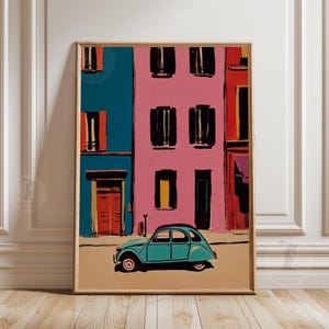 May include: A framed art print depicts a colorful street scene. Buildings in teal, pink, and red feature black window shutters and doors. A teal vintage car is parked on the tan street. The artwork is in a gold frame.
