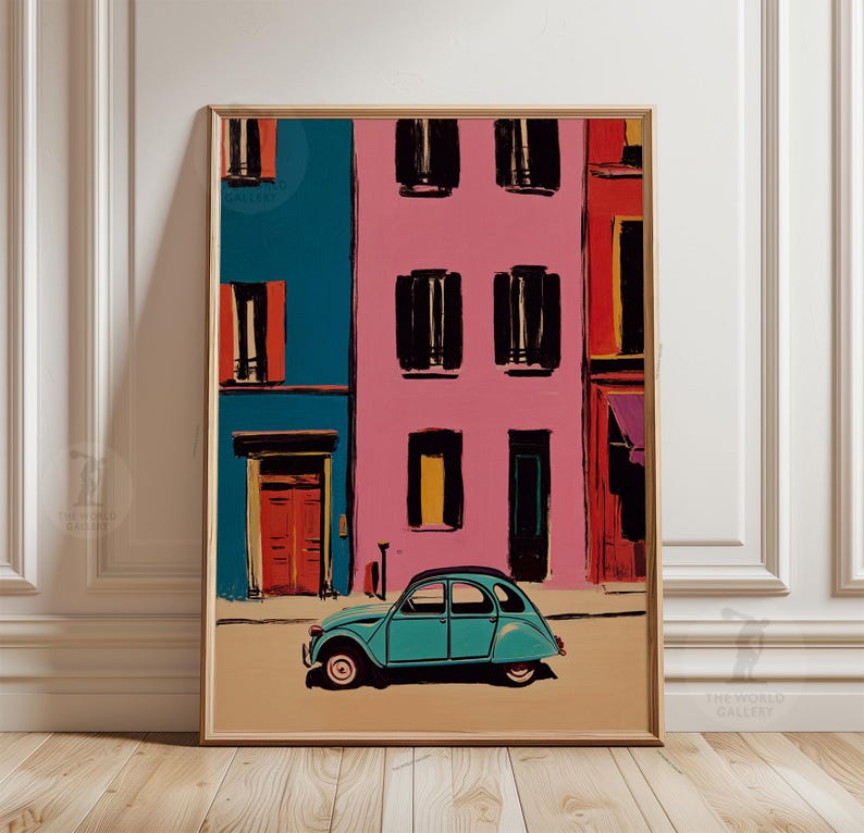 May include: A framed art print depicts a colorful street scene. Buildings in teal, pink, and red feature black window shutters and doors. A teal vintage car is parked on the tan street. The artwork is in a gold frame.