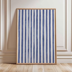 May include: A framed art print with vertical blue and white stripes. The artwork is presented in a wooden frame and leans against a white wall and wooden floor. The design evokes a coastal or nautical aesthetic.
