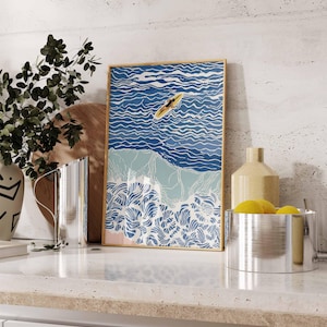 May include: A framed art print depicts a person on a yellow surfboard in blue ocean waves. The artwork has a gold frame and features a beach scene with white, blue, and light blue colors. The print is displayed on a white countertop.
